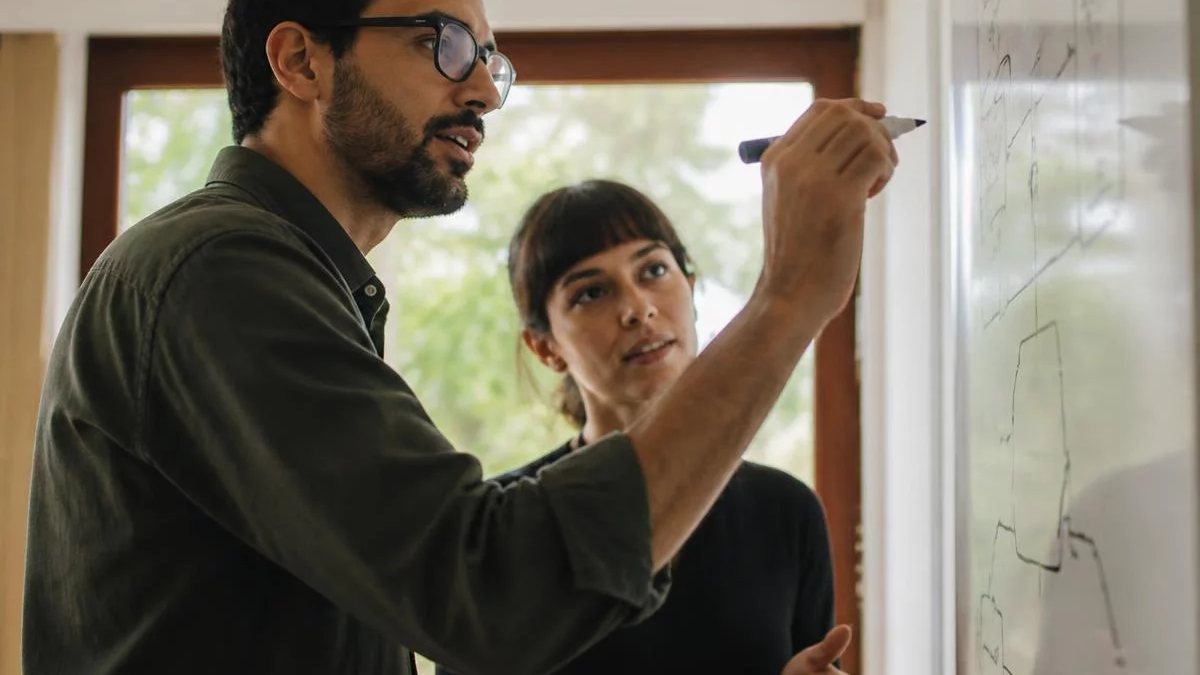 Duas participantes de pé diante do quadro branco, montando um argumento a caneta
